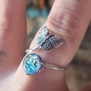 Side-angle close-up of a sterling silver butterfly cremation ring featuring a detailed butterfly accent and a round blue-green glass centerpiece containing cremation ash, shown worn on a finger with a softly blurred background.