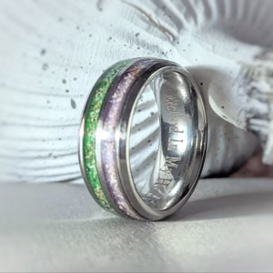 Dual Channel Cremation Ring with green and soft purple memorial glass inlay, shown resting on a white seashell tabletop surface