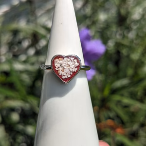 Memorial heart cremation ring displayed on a white ring cone outdoors, featuring a heart-shaped setting with cremation ash suspended in deep red resin on a silver band