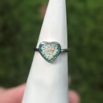Memorial heart cremation ring displayed on a white ring cone, featuring a heart-shaped setting with cremation ash suspended in teal resin on a silver band