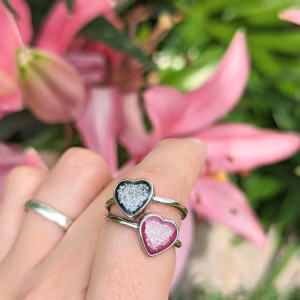 Memorial heart cremation rings worn on a hand outdoors, featuring two heart-shaped settings with cremation ash suspended in black and deep red resin on silver bands