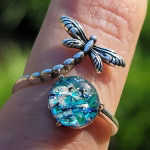 Close-up of the Dragonfly Cremation Ring in the Aqua Crinkle color variation, featuring shimmering aqua memorial glass and a detailed silver dragonfly resting on the ring band in bright natural sunlight