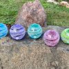 Five Infinite Staircase cremation paperweights displayed outdoors on a large stone, each featuring swirling glass bands with suspended cremation ashes. Colors shown from left to right are Blue, Amethyst, Aquamarine, Pink, and Granny Apple Green. A grassy yard and natural rocks appear softly in the background.
