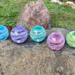 Five Infinite Staircase cremation paperweights displayed outdoors on a large stone, each featuring swirling glass bands with suspended cremation ashes. Colors shown from left to right are Blue, Amethyst, Aquamarine, Pink, and Granny Apple Green. A grassy yard and natural rocks appear softly in the background.