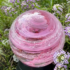 A hand-blown Infinite Staircase cremation paperweight in Opal Bright Pink, featuring the loved one’s ashes suspended in swirling pink glass ribbons. The sphere is photographed outdoors among small purple and white flowers.