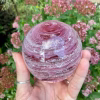 A hand-blown Infinite Staircase cremation paperweight in Opal Bright Pink glass, with the loved one’s ashes suspended in soft swirling pink ribbons. The sphere is held in a hand outdoors with blooming pink flowers in the background.