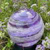 A hand-blown Infinite Staircase cremation paperweight in Amethyst glass, with suspended ashes swirling in layered lavender and purple ribbons. The sphere is photographed outdoors among delicate purple flowers.
