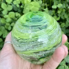 A hand-blown Infinite Staircase cremation paperweight made in Granny Apple Green glass, with ashes suspended in swirling ribbons throughout the sphere. It is shown resting in a hand against a backdrop of fresh green clover.