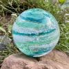 A hand-blown Infinite Staircase cremation paperweight featuring swirling bands of Aquamarine and Granny Apple Green glass with suspended ashes. The sphere rests on a natural stone outdoors, surrounded by soft greenery and small purple wildflowers.