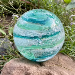 A hand-blown Infinite Staircase cremation paperweight featuring swirling bands of Aquamarine and Granny Apple Green glass with suspended ashes. The sphere rests on a natural stone outdoors, surrounded by soft greenery and small purple wildflowers.