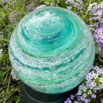 A Bristol Green and Aquamarine Infinite Staircase cremation paperweight shown outdoors among small purple flowers. The glass sphere features swirling layers of green tones with suspended cremation ashes and tiny bubbles, forming a spiral design that glows in natural light.