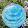 Close-up of an Infinite Staircase cremation paperweight in aquamarine glass, featuring swirling embedded ashes and delicate bubbles throughout. The memorial sphere rests among small purple flowers, with sunlight highlighting its vibrant blue color and spiral design.