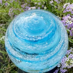 Close-up of an Infinite Staircase cremation paperweight in aquamarine glass, featuring swirling embedded ashes and delicate bubbles throughout. The memorial sphere rests among small purple flowers, with sunlight highlighting its vibrant blue color and spiral design.