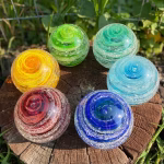 Top-down view of six Infinite Staircase cremation paperweights displayed on a tree stump in bright sunlight. The glass spheres feature swirling cremation ashes suspended within vivid colors: yellow, red, deep blue, sky blue, aquamarine, and granny apple green. Sunlight highlights the spiral design and sparkling details of each memorial piece.