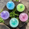 Top-down view of five Infinite Staircase cremation paperweights arranged in a circle on a weathered tree stump. Each glass sphere features swirling pastel colors with suspended cremation ashes. The colors shown are Blue, Granny Apple Green, Pink, Aquamarine, and Amethyst. Small green succulents grow through the center of the stump, adding a natural backdrop.