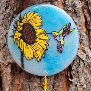 Round glass suncatcher featuring a sunflower and a hummingbird against a blue background, incorporating cremation ash, hanging outdoors against tree bark.