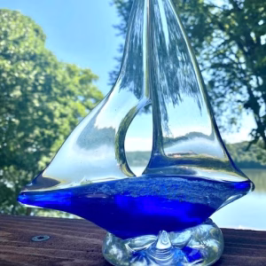 A hand-sculpted clear glass sailboat featuring a deep cobalt-blue glass base infused with cremation ash, shown outdoors with trees and water reflected through the sails.