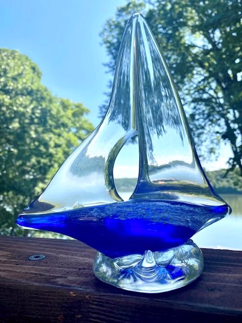 A hand-sculpted clear glass sailboat featuring a deep cobalt-blue glass base infused with cremation ash, shown outdoors with trees and water reflected through the sails.