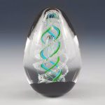 Front-facing view of a spiral egg-shaped cremation glass keepsake featuring a clear glass form with layered aqua and green spiral detailing and visible cremation ashes suspended inside.
