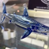 Side view of a glass shark memorial sculpture crafted in deep blue and clear glass, with cremation ash visibly suspended throughout the body, displayed on a glass shelf.
