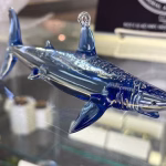 Side view of a glass shark memorial sculpture crafted in deep blue and clear glass, with cremation ash visibly suspended throughout the body, displayed on a glass shelf.