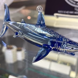 Side view of a glass shark memorial sculpture crafted in deep blue and clear glass, with cremation ash visibly suspended throughout the body, displayed on a glass shelf.