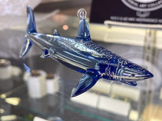 Side view of a glass shark memorial sculpture crafted in deep blue and clear glass, with cremation ash visibly suspended throughout the body, displayed on a glass shelf.