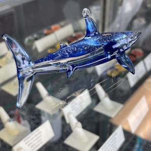 Glass shark memorial sculpture displayed on a glass surface, featuring deep blue and clear glass with visible cremation ash suspended inside the body.