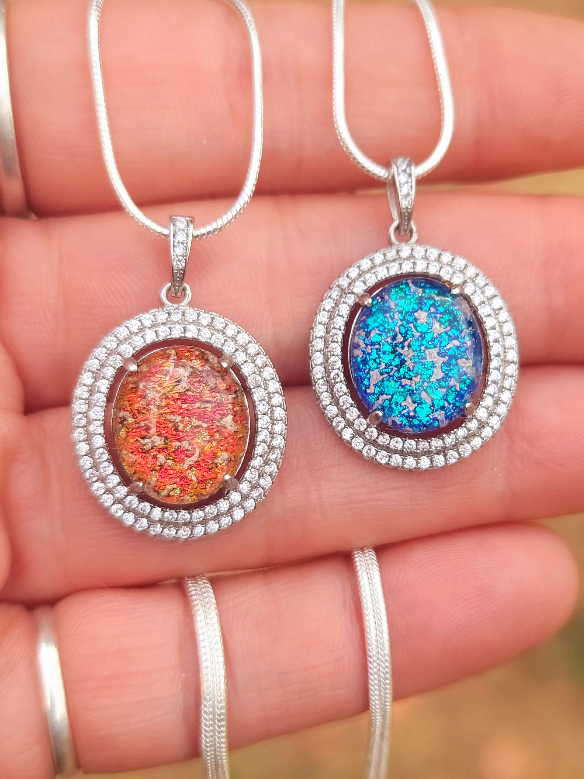 Halo Cremation Pendant with Cubic Zirconia in sterling silver, shown in blue and red orange memorial ash inlays on silver chains displayed side by side on a hand