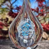 Clear teardrop memorial glass keepsake containing cremation ash, featuring a suspended inner teardrop with white ash and an aqua and blue spiral, photographed outdoors on stone with soft-focus red foliage and blue sky in the background.