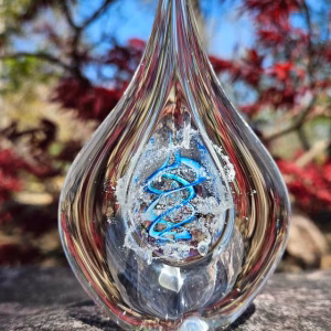 Clear teardrop memorial glass keepsake containing cremation ash, featuring a suspended inner teardrop with white ash and an aqua and blue spiral, photographed outdoors on stone with soft-focus red foliage and blue sky in the background.