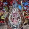 Clear teardrop memorial glass keepsake containing cremation ash, featuring a suspended inner teardrop with soft white ash and a flowing purple-pink spiral, photographed outdoors on stone with blurred red foliage in the background.