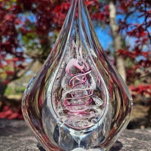 Clear teardrop memorial glass keepsake containing cremation ash, featuring a suspended inner teardrop with soft white ash and a flowing purple-pink spiral, photographed outdoors on stone with blurred red foliage in the background.
