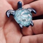 Handcrafted glass sea turtle cremation pendant with deep blue flippers and a textured aqua shell containing white flecks of encased cremation ashes, shown resting in the hand for a detailed close-up view