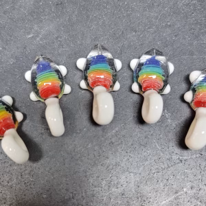 Five small glass turtle pendants arranged in a row on a work surface, each featuring a rainbow-colored shell with cremation ash suspended inside and white glass limbs and tail.