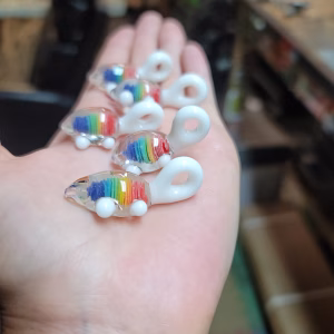 Hand holding several small glass rainbow turtle pendants, each featuring a vibrant rainbow-colored shell with cremation ash encased inside and smooth white glass limbs and tail.