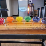 Six glass memorial paperweights with cremation ash displayed in a row on a wooden workbench, arranged in a rainbow spectrum—from red, orange, yellow, green, blue, to purple—each featuring swirling color bands and suspended ash within the clear glass.