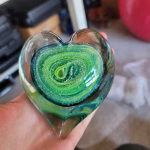 Top view of a clear glass heart-shaped paperweight held in a hand, showcasing layered green and teal swirling bands encased inside, with cremation ash forming delicate white speckled trails around the spiral design.