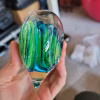 Side view of a clear glass heart-shaped paperweight held in a hand, featuring layered green and blue swirling bands encased inside, with cremation ash forming fine white trails throughout the design.
