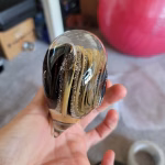 Side view of a clear glass heart-shaped paperweight held in a hand, featuring layered black, tan, and cream swirling bands inside, with fine cremation ash trails suspended throughout the design.