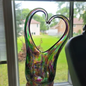 Handblown glass heart memorial sculpture with cremation ashes, featuring an iridescent multicolor finish and open heart shape, held in front of a window with natural light shining through