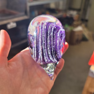 Side view of a clear glass heart-shaped paperweight held in a hand, featuring vibrant purple swirling bands encased inside, with cremation ash forming shimmering white trails throughout the design.