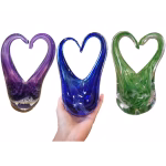 Three glass heart-shaped cremation sculptures displayed side-by-side in purple, blue, and green color variations. Each sculpture features flowing glass curves forming an open heart shape with cremation ash swirls suspended inside the base; the center blue sculpture is shown being held in a hand.