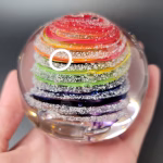 Hand holding a round glass paperweight featuring vibrant rainbow-colored swirling bands encased inside, with delicate cremation ash patterns suspended throughout the clear glass.