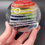 Hand holding a round glass paperweight featuring vibrant rainbow-colored swirling bands encased inside, with delicate cremation ash patterns suspended throughout the clear glass.
