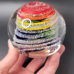 Hand holding a round glass paperweight featuring vibrant rainbow-colored swirling bands encased inside, with delicate cremation ash patterns suspended throughout the clear glass.