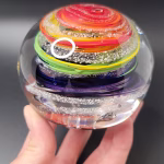 Hand holding a round glass paperweight featuring vibrant rainbow-colored swirling bands encased inside, with delicate cremation ash patterns suspended throughout the clear glass.
