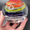 Hand holding a round glass paperweight featuring vibrant rainbow-colored swirling bands encased inside, with delicate cremation ash patterns suspended throughout the clear glass.