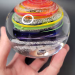 Hand holding a round glass paperweight featuring vibrant rainbow-colored swirling bands encased inside, with delicate cremation ash patterns suspended throughout the clear glass.