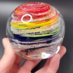 Hand holding a round glass paperweight featuring vibrant rainbow-colored swirling bands encased inside, with delicate cremation ash patterns suspended throughout the clear glass.